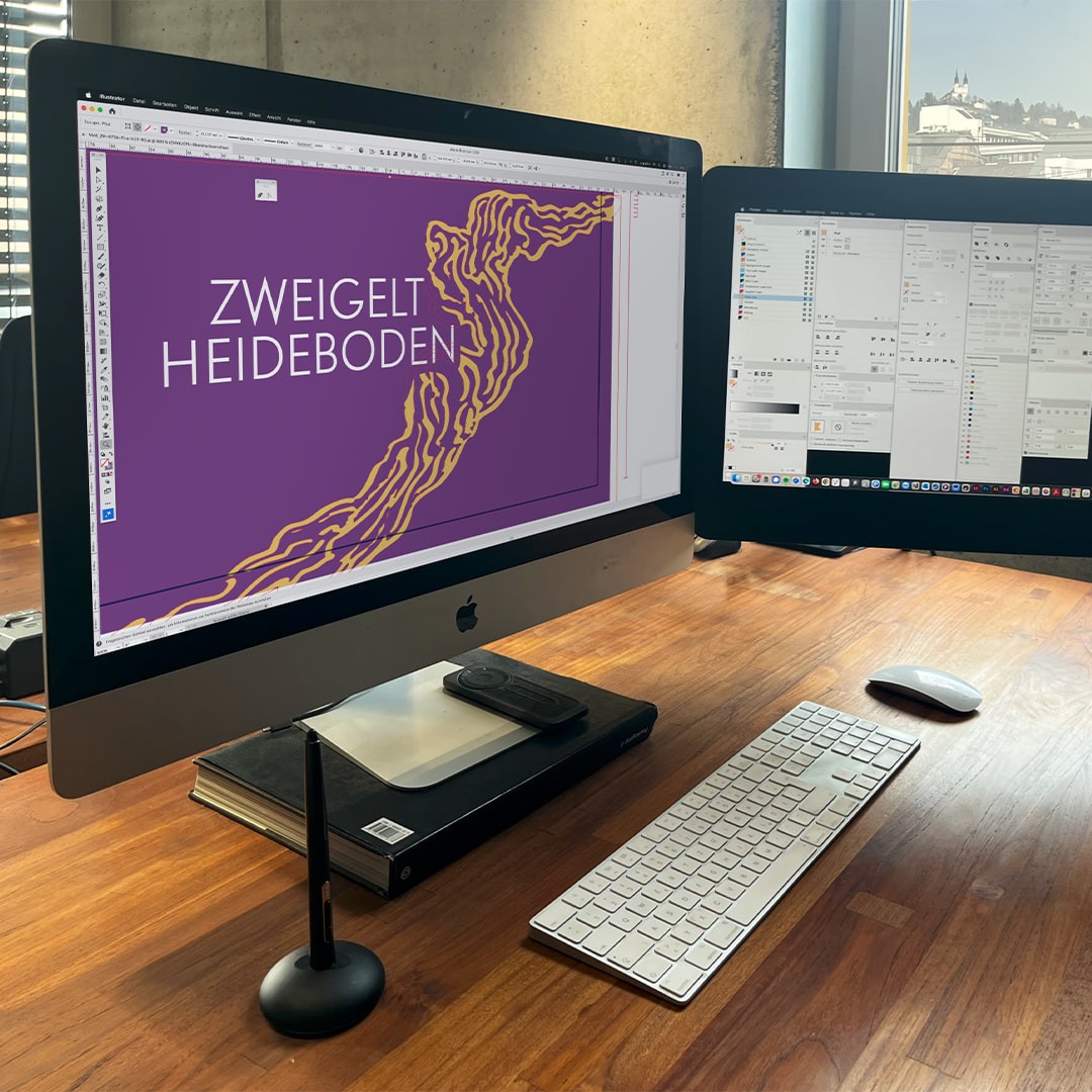Arbeitsplatz mit zwei Bildschirmen und digitalem Entwurf für das Vinion Edition Zweigelt Heideboden Etikett in Lila und Gold.