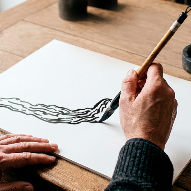 Hand zeichnet mit Pinsel eine schwarze Linienillustration auf weissem Papier.